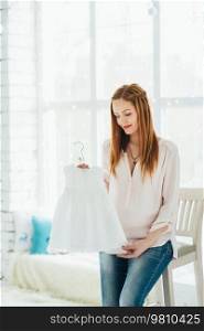 a red-haired pregnant girl in a light blouse and blue jeans sits on a high chair in a bright room
