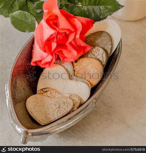 A red rose lying on top of a silver bowl full of porcelain hearts with doily textures inprinted on them.