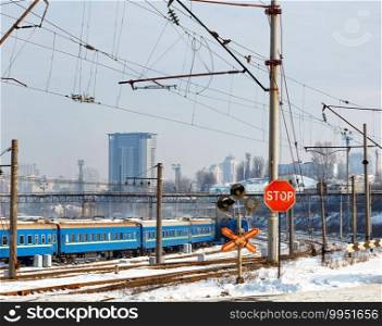 A red stop sign at a railroad crossing against the backdrop of railroad tracks, blue train carriages and cityscape at the entrance to a winter city.. Red stop sign at the railway crossing against the background of the railway station and buildings of the winter city.