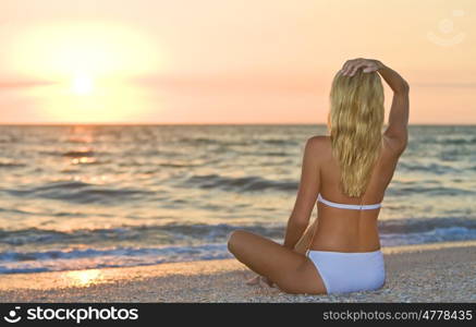 A relaxed sexy young blonde woman or girl wearing a bikini sitting on a deserted tropical beach at sunset or sunrise