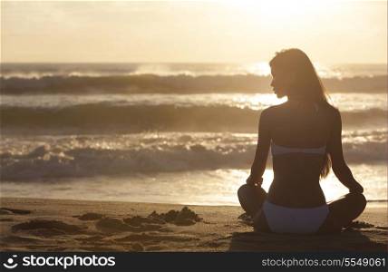 A relaxed sexy young brunette woman or girl wearing a bikini sitting on a deserted tropical beach at sunset or sunrise