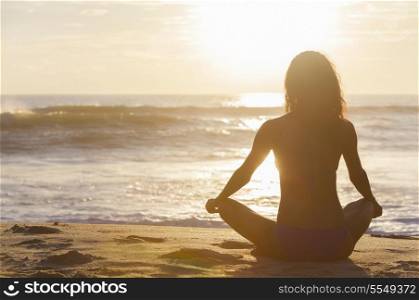 A relaxed sexy young brunette woman or girl wearing a bikini sitting on a deserted tropical beach at sunset or sunrise