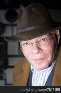 A rich contrast close up portrait of a senior man, slightly leaning forward, wearing a hat and overcoat against a grungy looking industrial background