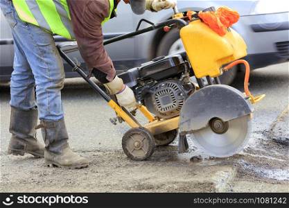A road maintenance officer in a green reflective vest starts the engine of the gasoline cutter to remove the old asphalt during repairs on the roadway.. A worker in a green reflective vest starts the engine of a gasoline cutter to cut and clean bad asphalt on the roadway.