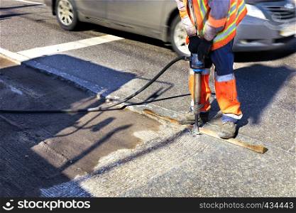 A road maintenance worker removes old asphalt on the roadway with a jackhammer into an excavator during road construction.. A worker clears a piece of asphalt with a pneumatic jackhammer in road construction.