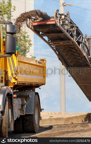 A road milling machine removes the top layer of old asphalt from the road and loads it onto a dump truck on a conveyor belt on a summer day. Copy space. Vertical image.. A road router removes the top layer of old asphalt from a section of road and loads it onto a tipper.