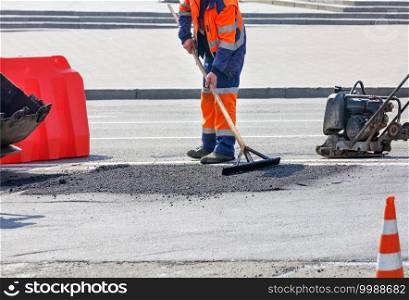 A road worker in a bright orange overalls on a fenced-off work section of the road patches a pothole with hot asphalt and r&s it with a petrol vibratory plate.. A road worker in a bright orange overalls levels fresh asphalt with a wooden level for later compaction.