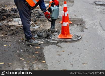 A road worker in a reflective orange vest smashes the asphalt with an electric jackhammer around a manhole on the roadway surrounded by red cones. Copy the space.. A road worker smashes asphalt with an electric jackhammer around a sewer hatch on the road.