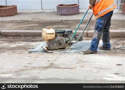 A road worker in an orange vest patches a pothole in an old road repairing it with fresh asphalt using an old petrol vibrating plate compactor. Copy space.. A road worker is patching up a pothole in an old road using an old petrol plate compactor.