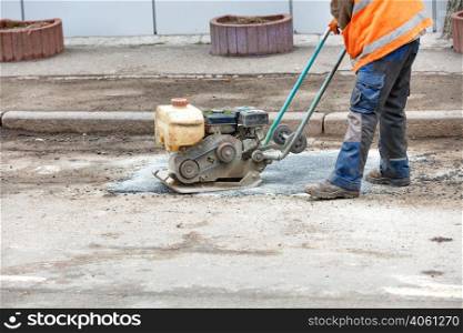 A road worker in an orange vest patches a pothole in an old road repairing it with fresh asphalt using an old petrol vibrating plate compactor. Copy space.. A road worker is patching up a pothole in an old road using an old petrol plate compactor.