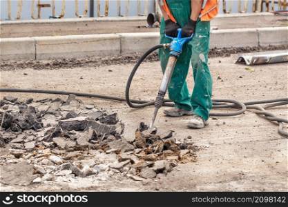 A road worker in green overalls loosens old asphalt with a two-handed pneumatic jackhammer at a road workplace on a summer day. Copy space.. A worker actively loosens old asphalt with a pneumatic jackhammer while repairing a section of a road.