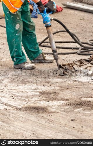 A road worker in green overalls smashes old asphalt with a pneumatic jackhammer on the road on a summer day. Vertical image. Copy space.. A road worker smashes old asphalt with a pneumatic jackhammer at a work site.