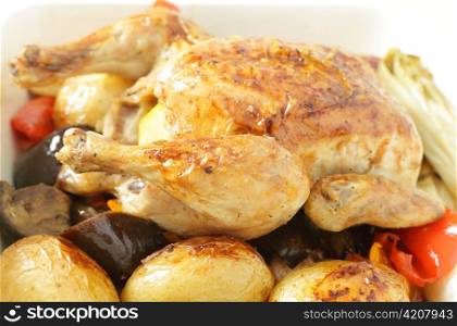 A roast chicken in a serving bowl on a bed of oven roasted vegetables, including potatoes, capsicum, endive, eggplant and garlic.