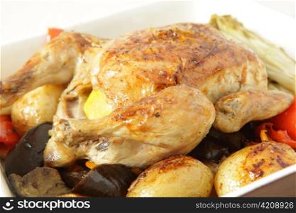 A roast chicken in a serving bowl on a bed of oven roasted vegetables, including potatoes, capsicum, endive, eggplant and garlic.