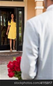A romantic African American man bringing flowers to his happy wife or girlfriend who is standing waiting for him at the door of their home.
