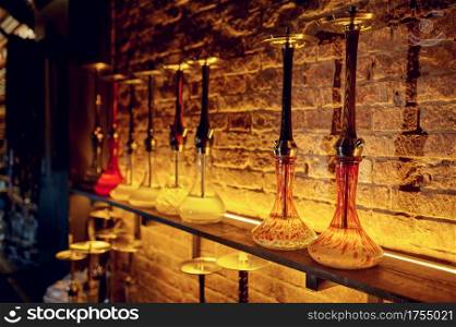 A row of hookahs with glass bulbs at the brick wall, nobody. Shisha bar equipment, traditional smoke culture, tobacco aroma. A row of hookahs with glass bulbs, nobody