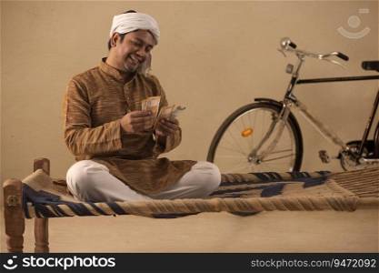 A RURAL MAN SITTING ON A COT AND HAPPILY COUNTING MONEY