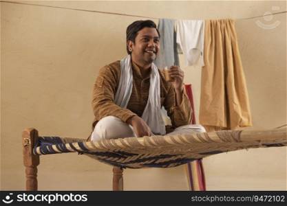 A RURAL MAN SITTING ON A COT HOLDING A GLASS OF TEA