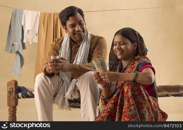 A RURAL WIFE HAPPILY COUNTING MONEY WHILE HUSBAND DRINKS TEA