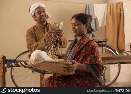 A RURAL WOMAN SITTING AND HUSKING RICE GRAINS WHILE HUSBAND COUNTS MONEY