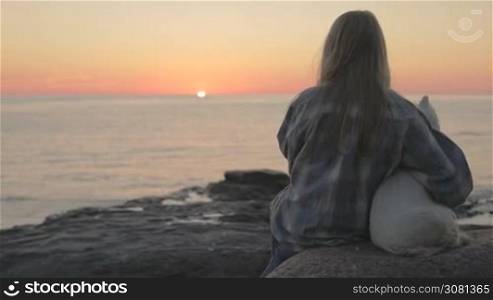 A sad little girl with a dog is sitting on the sea coast on the stones and watching the sunset. Slow motion
