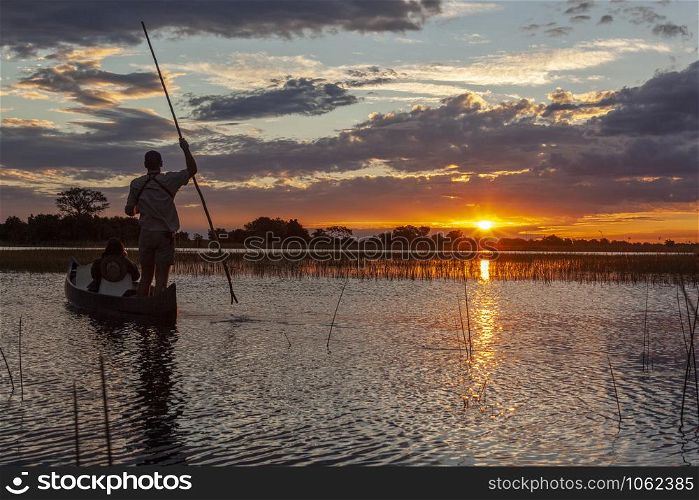 A safari guide with a tourist in a Makoro (dugout canoe) at sunset in the Okavango Delta in northern Botswana, Africa.