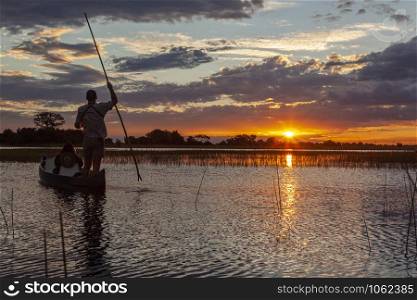 A safari guide with a tourist in a Makoro (dugout canoe) at sunset in the Okavango Delta in northern Botswana, Africa.