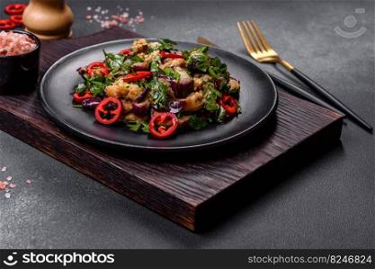 A salad of baked aubergine, sweet pepper, garlic, zucchini and parsley in a black plate against a dark concrete background. Vegetarian food. A salad of baked aubergine, sweet pepper, garlic, zucchini and parsley in a black plate