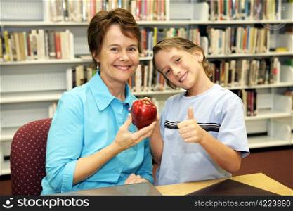A school boy giving an apple to his teacher and a thumbsup sign to the camera.