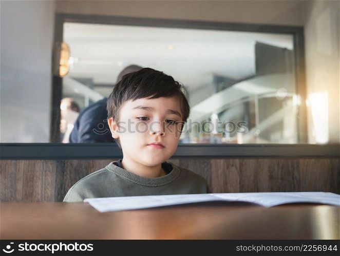 A School kid reading or choosing food in the menu ready to order food for lunch, Child boy sitting and waiting for his meal in the restaurant.
