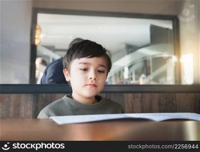 A School kid reading or choosing food in the menu ready to order food for lunch, Child boy sitting and waiting for his meal in the restaurant.