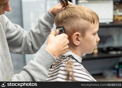 A schoolboy is sitting in a barbershop, doing his hair with a clipper. A boy is sitting in a barber shop, cutting his hair with a clipper