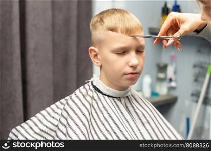 A schoolboy sits with his eyes closed in a barbershop, doing his hair with scissors for haircuts. A schoolboy is sitting in a barbershop, doing his hair with scissors for haircuts
