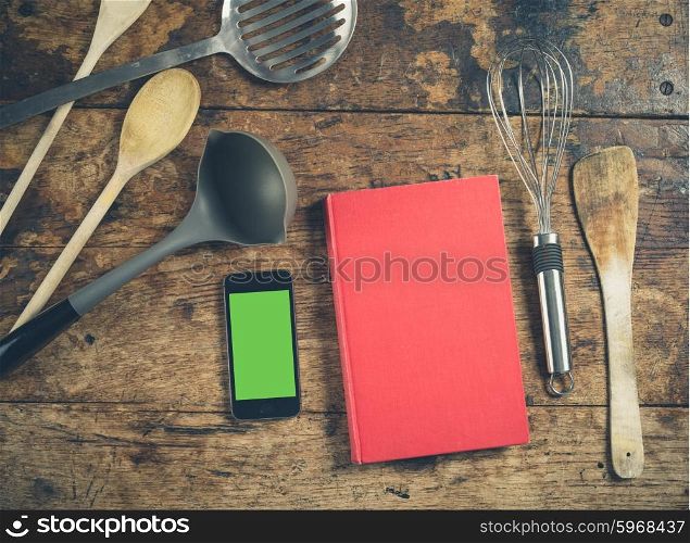 A selection of kitchen utensils on a wooden table with a book and a smart phone with a green screen chromakey