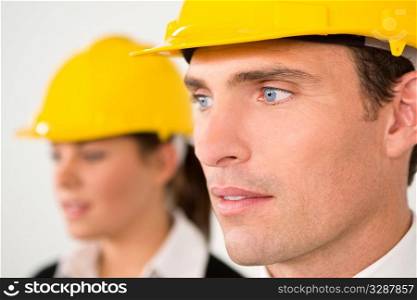 A selective focus industrial concept shot showing a man and woman dressed in hard hats. The focus is on the man in the foreground