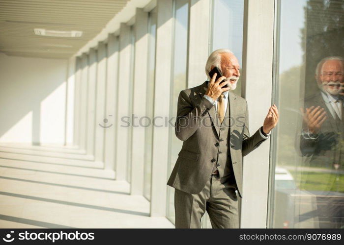 A senior business man stands in an office hallway, focused on his mobile phone. He is dressed in formal attire, exuding confidence and professionalism