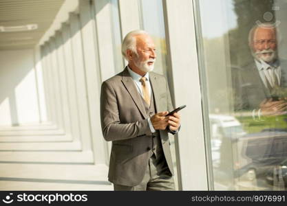 A senior business man stands in an office hallway, focused on his mobile phone. He is dressed in formal attire, exuding confidence and professionalism