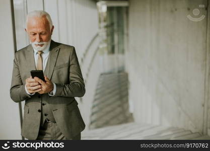 A senior business man stands in an office hallway, focused on his mobile phone. He is dressed in formal attire, exuding confidence and professionalism