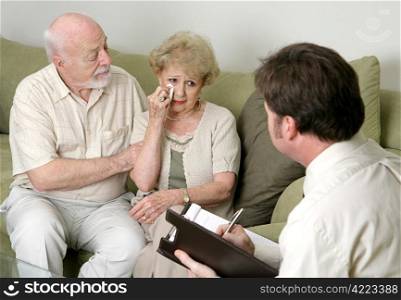 A senior couple in counseling - either grief counseling or marriage counseling. The wife is crying and the husband is wiping her tears and trying to console her.