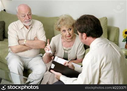 A senior couple in marriage counseling. She&rsquo;s complaining to the therapist about her husband while he looks on in disbelief. Focus on wife.