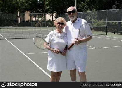 A senior couple on the tennis courts wearing sunglasses and getting ready for a game.