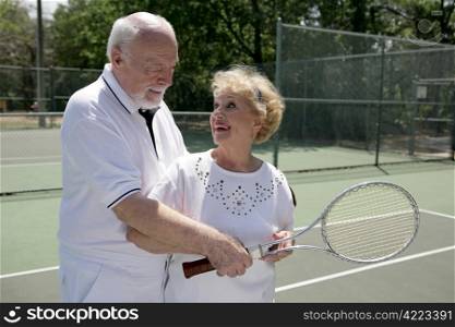 A senior couple playing tennis together. He&rsquo;s showing her how to grip her racquet.