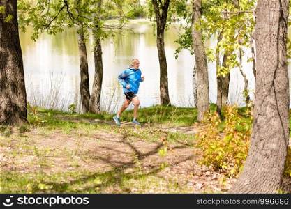 A senior man dressed in black and blue is running close to the lake during a warm spring day