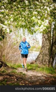 A senior man dressed in black and blue is running in the forest, close to the lake, under an apple blossom, during a warm spring day