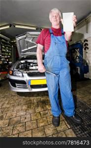 A senior motor mechanic, holding up a blank cart with a wrench in his hands, posing in front of the car he&rsquo;s been working on