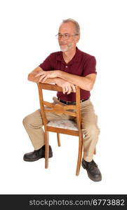 A serious older man in beige pants and burgundy t-shirt sitting backwardson a chair, isolated for white background.