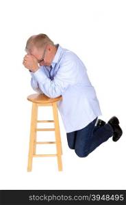 A serious senior kneeling on a chair praying with folded hands in a blueshirt and jeans, isolated for white background.