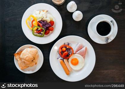 A set of American breakfast sunny side up eggs, sausage, ham, fresh salad bell peppers, tomato, croissant bread with black coffee. Top view