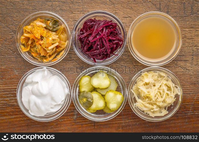 a set of fermented food great for gut health - top view of glass bowls against rustic wood: kimchi, red beets, apple cider vinegar, coconut milk yogurt, cucumber pickles, sauerkraut
