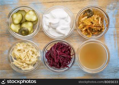 a set of fermented food great for gut health - top view of glass bowls against grunge wood: cucumber pickles, coconut milk yogurt, kimchi, sauerkraut, red beets, apple cider vinegar